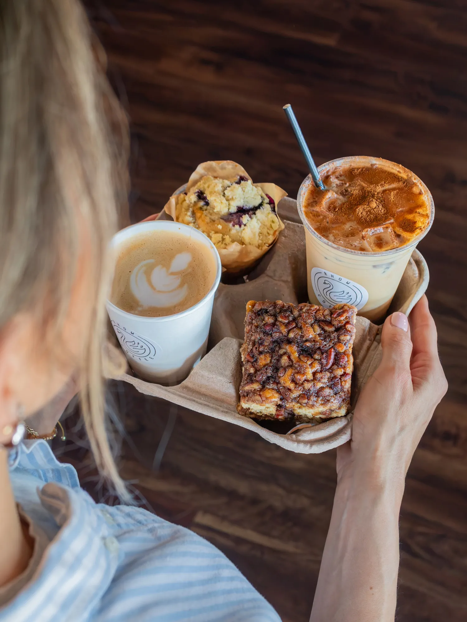 Hands holding a RHOW Coffee tray with a latte, an iced coffee, a muffin, and a pastry bar