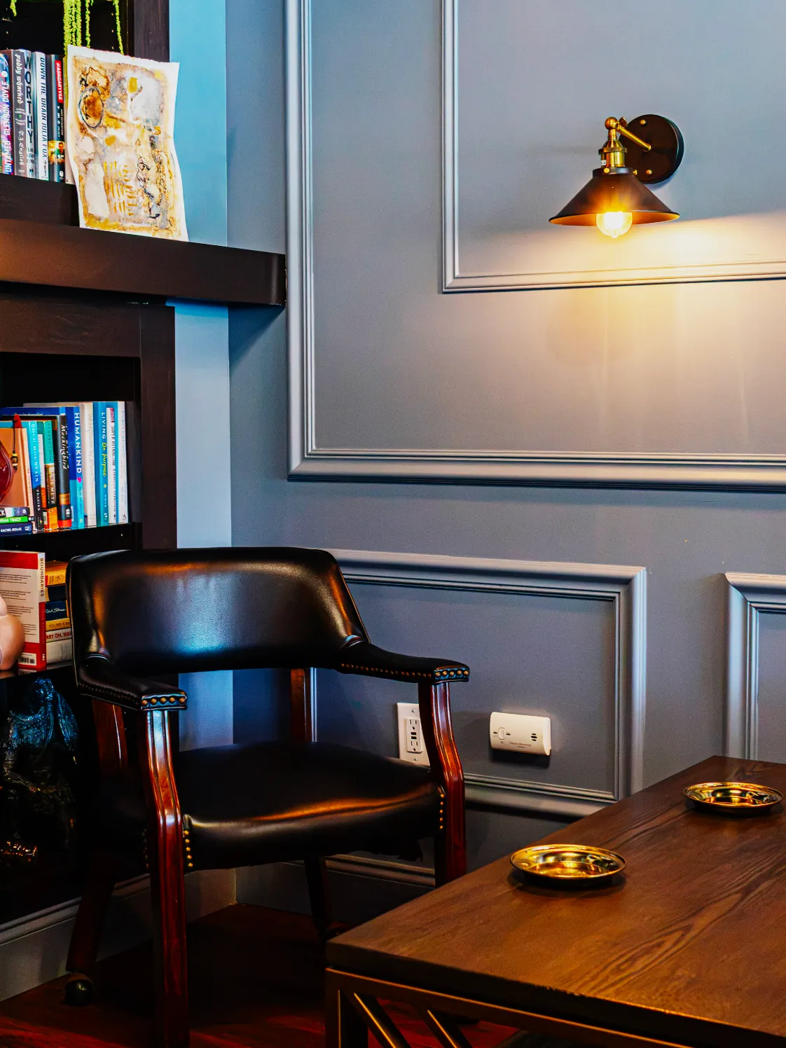 Leather armchair and wooden table beside bookshelves under a warm wall sconce