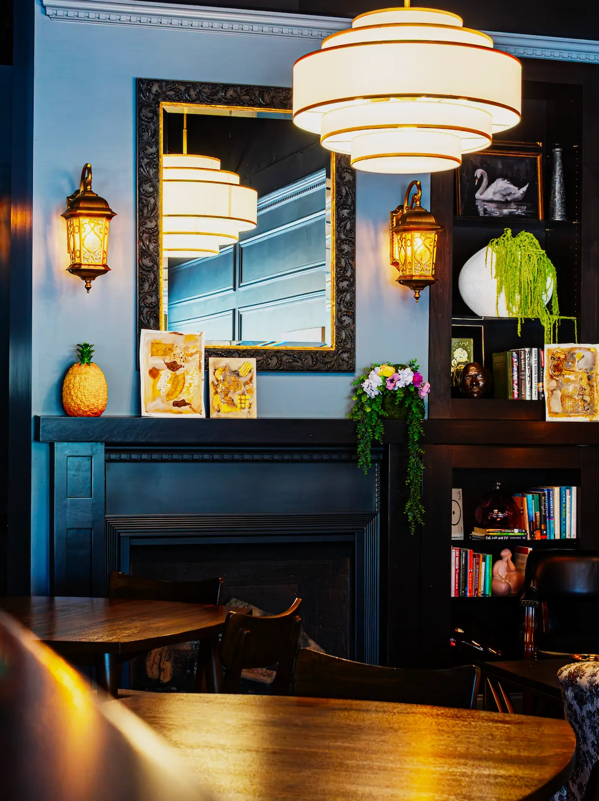 Shop interior with a gilded mirror over a dark fireplace, a pendant light, bookshelves, and plants